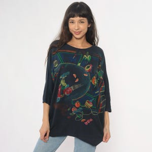 May include: Dark navy blue oversized t-shirt with a colorful abstract design. The graphic features a scooter, fruit, and other shapes in red, green, and orange. The shirt has three-quarter sleeves and a round neckline.