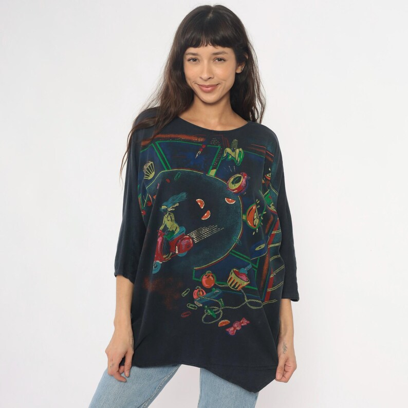 May include: Dark navy blue oversized t-shirt with a colorful abstract design. The graphic features a scooter, fruit, and other shapes in red, green, and orange. The shirt has three-quarter sleeves and a round neckline.