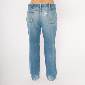 May include: Light blue denim jeans with a classic fit. The jeans feature a faded wash, distressed details, and the Wrangler logo on the back pockets. The jeans have a straight leg and a worn hem.