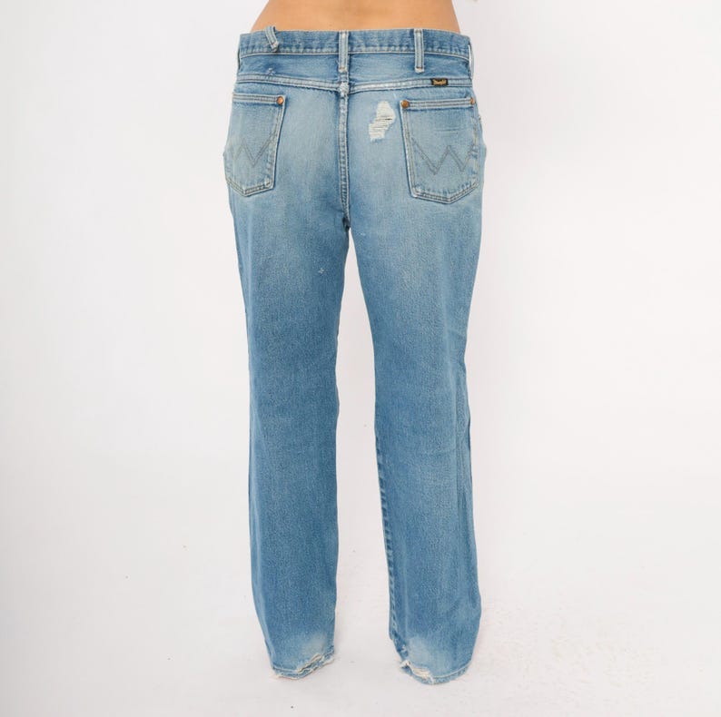 May include: Light blue denim jeans with a classic fit. The jeans feature a faded wash, distressed details, and the Wrangler logo on the back pockets. The jeans have a straight leg and a worn hem.