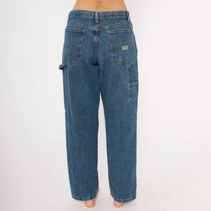 May include: Blue denim carpenter jeans with multiple pockets, including a hammer loop and a patch on the back. The jeans have a relaxed fit and a classic five-pocket design. The jeans are made of a durable denim material.