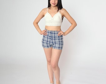 May include: A woman wearing a white crochet top and blue and white plaid shorts. She is standing with her hands on her hips and is wearing white sneakers.