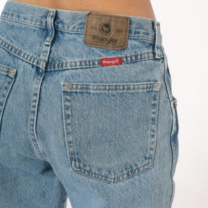 May include: Light blue denim jeans with a classic five-pocket design. The back features a rectangular pocket and a brown leather patch with the "Wrangler" logo. A red "Wrangler" tag is sewn on the pocket. The jeans are made of durable denim.
