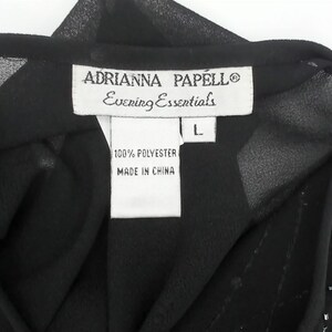 May include: Close-up of a black evening top with a label that reads "ADRIANNA PAPELL Evening Essentials". The label also indicates the size is L and the fabric is 100% polyester, made in China.