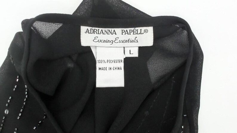 May include: Close-up of a black evening top with a label that reads "ADRIANNA PAPELL Evening Essentials". The label also indicates the size is L and the fabric is 100% polyester, made in China.