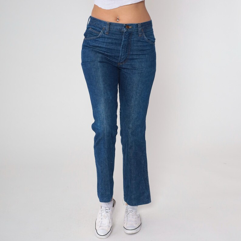 May include: Dark blue denim jeans with a classic five-pocket design. The jeans have a straight leg fit and a button and zipper closure. The model is wearing white sneakers.