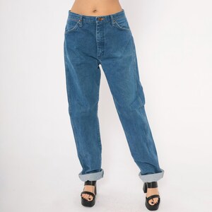 May include: Blue denim jeans with a button and zipper closure. The jeans have a classic five-pocket design and are cuffed at the ankles. The jeans are worn with black platform sandals.