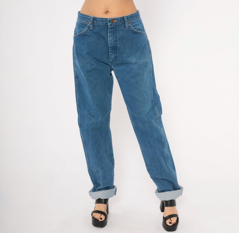 May include: Blue denim jeans with a button and zipper closure. The jeans have a classic five-pocket design and are cuffed at the ankles. The jeans are worn with black platform sandals.