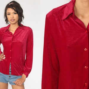 May include: A long-sleeved button-down shirt in a deep red velvet fabric. The shirt features a classic collar, a front button placket with matching red buttons, and gathered detailing at the shoulder seams, providing a structured yet soft appearance.