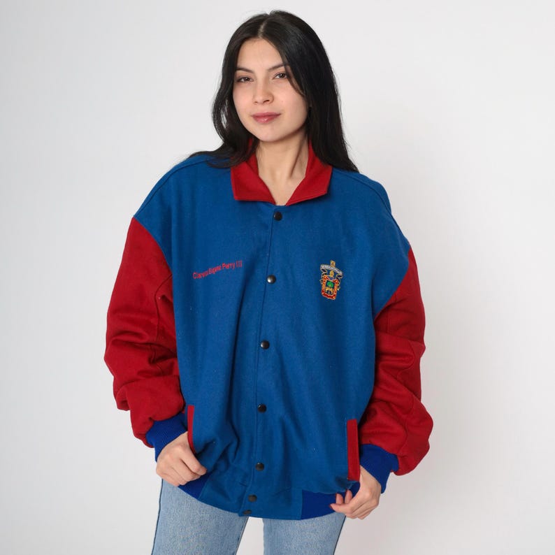 May include: A blue and red varsity jacket with a red embroidered crest on the left side. The jacket has a snap closure and a ribbed collar, cuffs, and hem.