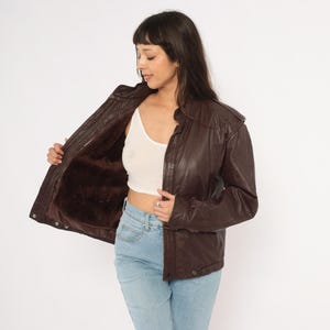 May include: A dark brown leather jacket with a fur lining. The jacket has a zip-up front, a collar, and pockets. The model is wearing a white tank top and blue jeans.