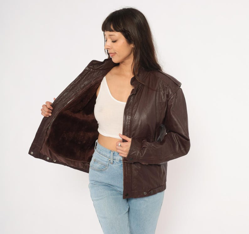 May include: A dark brown leather jacket with a fur lining. The jacket has a zip-up front, a collar, and pockets. The model is wearing a white tank top and blue jeans.