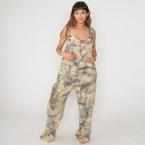 May include: Full-length view of camouflage overalls. The overalls feature a pattern of brown, green, and tan tree branches and leaves. The bib overalls have adjustable straps and a button closure. The garment is made of a durable fabric.
