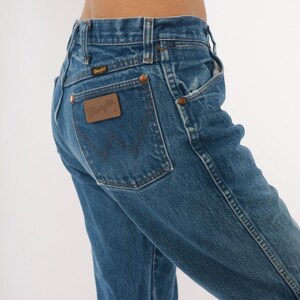 Pode incluir: Jeans Wrangler de ganga azul. Os jeans t&ecirc;m um design cl&aacute;ssico de cinco bolsos, com rebites de cobre e um patch de couro castanho no bolso traseiro. O logotipo Wrangler &eacute; vis&iacute;vel no patch. Os jeans s&atilde;o de lavagem m&eacute;dia e parecem vintage.