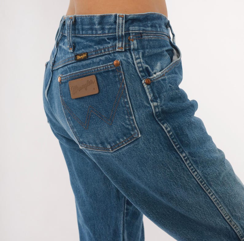 Pode incluir: Jeans Wrangler de ganga azul. Os jeans t&ecirc;m um design cl&aacute;ssico de cinco bolsos, com rebites de cobre e um patch de couro castanho no bolso traseiro. O logotipo Wrangler &eacute; vis&iacute;vel no patch. Os jeans s&atilde;o de lavagem m&eacute;dia e parecem vintage.