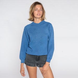 May include: A blue long-sleeve sweatshirt with a crew neck. The sweatshirt has a slightly faded appearance and is paired with black denim shorts. The model is standing in front of a white background.