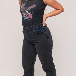 May include: Black tank top with a graphic of an eagle, roses, and a motorcycle. The tank top has a scalloped neckline. The model is wearing black denim jeans.