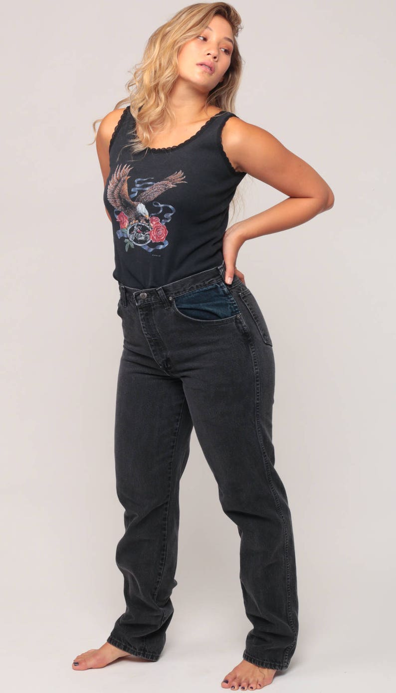 May include: Black tank top with a graphic of an eagle, roses, and a motorcycle. The tank top has a scalloped neckline. The model is wearing black denim jeans.