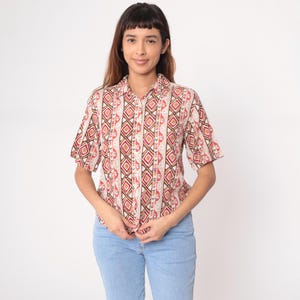 May include: A button-down shirt with a geometric pattern in shades of red, brown, and beige. The short-sleeved shirt is paired with light blue jeans. The shirt features a collared neckline and is buttoned up.