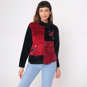 Puede incluir: Un jersey de cuello alto de patchwork en negro y rojo. El diseño incluye un cuadrado rojo con un estampado floral y un cuadrado rojo liso. Los cuadrados negros tienen diseños florales bordados. La parte superior de manga larga está hecha de un material suave y afelpado.