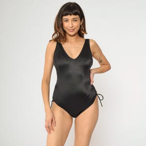 May include: A black one-piece swimsuit with a V-neck and adjustable straps. The swimsuit has a smooth, satin-like finish and a fitted silhouette.