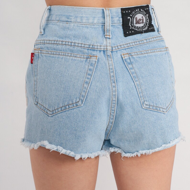 Puede incluir: Pantalones cortos de mezclilla azul claro con dobladillo deshilachado y un bolsillo trasero. Los pantalones cortos tienen una etiqueta en blanco y negro con el texto "Riding Wear Lei Quality Product" en el bolsillo trasero derecho.