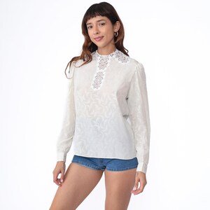 May include: White long-sleeve blouse with a lace collar and a textured pattern. The blouse has a high neckline and a relaxed fit.