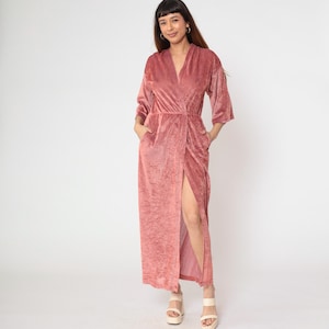 May include: A dusty rose velvet wrap dress with a V-neckline and three-quarter sleeves. The dress features a high slit and pockets. The model is wearing platform sandals. The dress is floor-length.