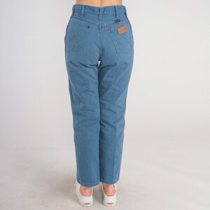 Op de afbeelding: Een paar blauwe denim jeans met een hoge taille en een rechte pijp. De jeans heeft een bruin leren patch op de achterkant met het Wrangler-logo.