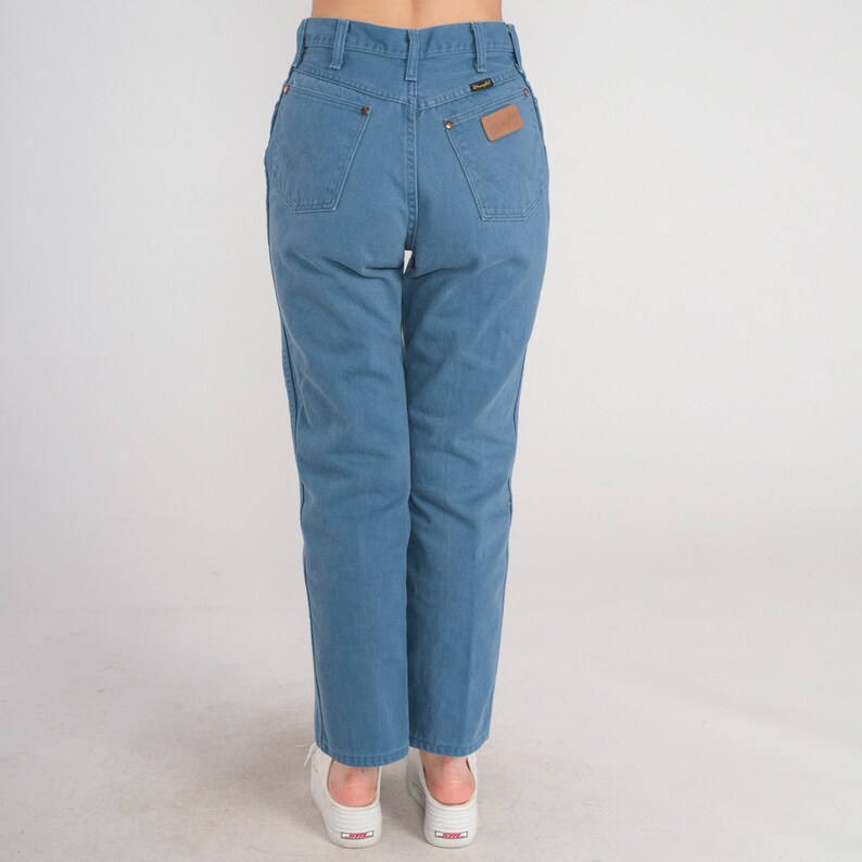 Op de afbeelding: Een paar blauwe denim jeans met een hoge taille en een rechte pijp. De jeans heeft een bruin leren patch op de achterkant met het Wrangler-logo.