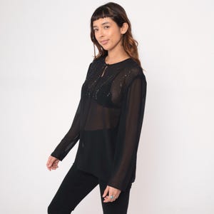 May include: Black sheer long-sleeve blouse with beaded embellishments around the neckline and a keyhole detail. The blouse is paired with black pants. The top is a semi-sheer material.