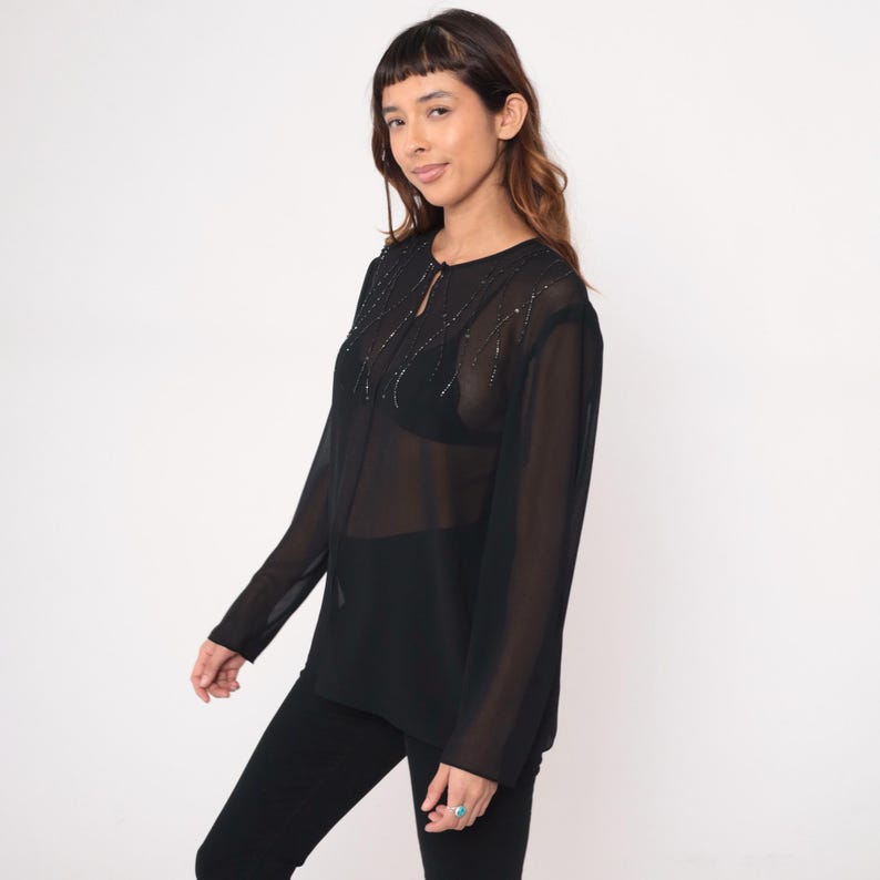 May include: Black sheer long-sleeve blouse with beaded embellishments around the neckline and a keyhole detail. The blouse is paired with black pants. The top is a semi-sheer material.