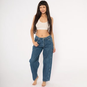 May include: A woman wearing a white crop top and blue denim carpenter jeans. The jeans have a relaxed fit and feature multiple pockets and a hammer loop. The model is barefoot, standing against a white background.