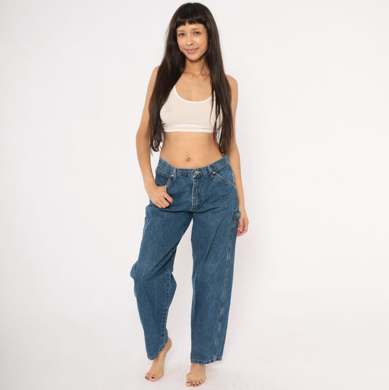 May include: A woman wearing a white crop top and blue denim carpenter jeans. The jeans have a relaxed fit and feature multiple pockets and a hammer loop. The model is barefoot, standing against a white background.