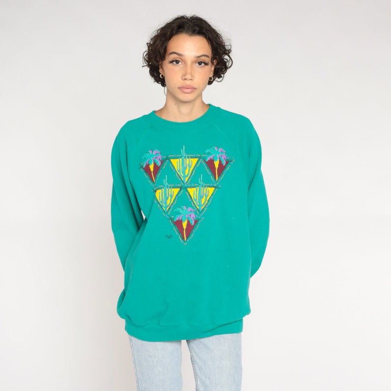 May include: Teal-colored sweatshirt with a triangular design featuring palm trees and cacti. The graphic has yellow, purple, and red accents. The long-sleeved top is a relaxed fit.