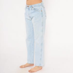May include: Light blue denim jeans with a classic five-pocket design. The jeans have a straight leg cut and a faded wash, giving them a vintage look. The jeans are worn by a person.