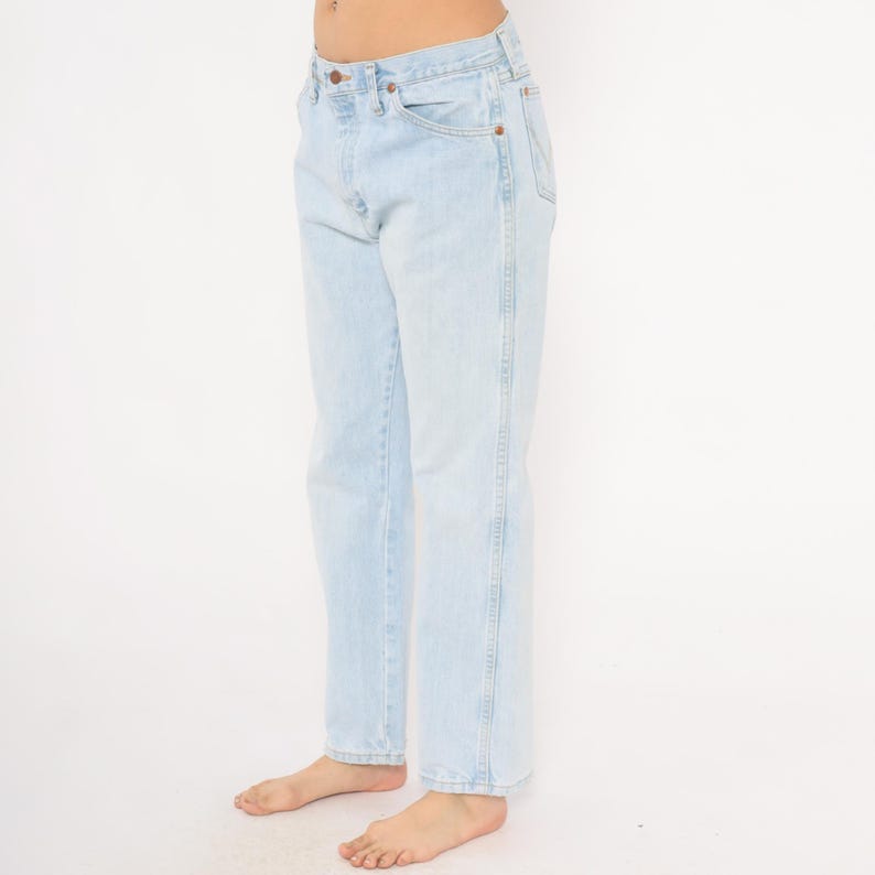 May include: Light blue denim jeans with a classic five-pocket design. The jeans have a straight leg cut and a faded wash, giving them a vintage look. The jeans are worn by a person.