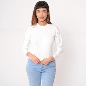 Puede incluir: Una camisa blanca de manga larga con cuello redondo y vaqueros azul claro. La camisa tiene un diseño moteado. La modelo lleva la camisa y los vaqueros.