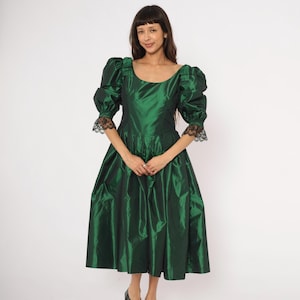 May include: A dark emerald green dress with a scoop neckline and puffed sleeves. The dress features a fitted bodice and a full skirt. The sleeves are accented with black lace trim. The dress is knee-length and made of a shiny fabric.
