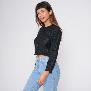 Puede incluir: Top negro de manga larga con un patr&oacute;n texturizado y botones dorados. El top est&aacute; abotonado y se usa con vaqueros azul claro. El top tiene cuello redondo y mangas largas.