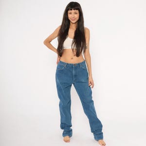 May include: Blue denim jeans with a classic straight leg fit. The jeans have a button and zipper closure and five-pocket styling. The denim is a medium wash, and the jeans appear to be vintage.