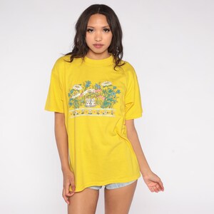 May include: A yellow t-shirt with a floral design featuring daisies and other flowers. The graphic includes a row of small, decorative elements at the bottom. The shirt has short sleeves and a crew neck.