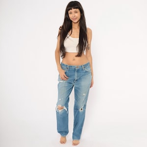 May include: Light blue denim jeans with distressed details, including ripped knees. The jeans have a classic five-pocket design and a button and zipper closure. The model is wearing a white crop top.