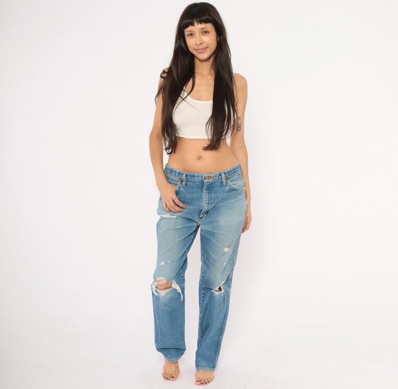 May include: Light blue denim jeans with distressed details, including ripped knees. The jeans have a classic five-pocket design and a button and zipper closure. The model is wearing a white crop top.