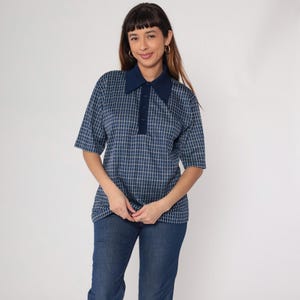 May include: A short-sleeved polo shirt with a navy blue collar and a blue and white grid pattern. The shirt has a three-button placket and is paired with blue jeans. The model is wearing gold hoop earrings.