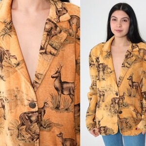 May include: A tan blazer with a repeating pattern of deer in a natural landscape. The blazer has a notched collar, two buttons, and long sleeves. The deer are depicted in shades of brown and black against a tan background.