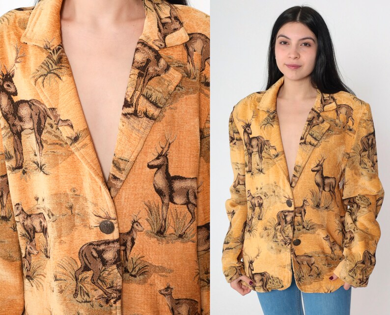 May include: A tan blazer with a repeating pattern of deer in a natural landscape. The blazer has a notched collar, two buttons, and long sleeves. The deer are depicted in shades of brown and black against a tan background.