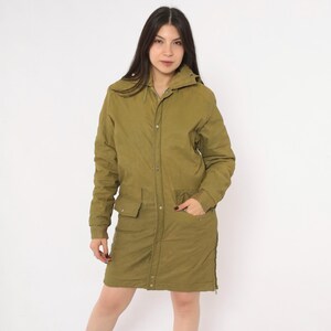 May include: An olive green long-length jacket featuring a hood, a front snap-button closure, long sleeves with ribbed cuffs, and two large front pockets with flaps. The jacket has a straight hemline and a solid, matte-textured fabric construction.