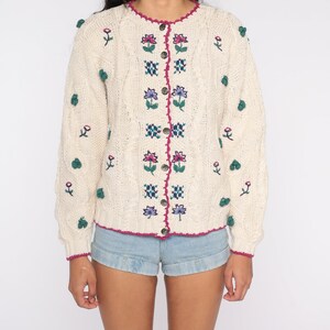 May include: A cream-colored knitted cardigan with a floral embroidered design. The cardigan has a button-up front and a scalloped neckline. The cardigan is decorated with small green pom-poms.