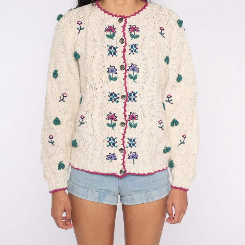 May include: A cream-colored knitted cardigan with a floral embroidered design. The cardigan has a button-up front and a scalloped neckline. The cardigan is decorated with small green pom-poms.